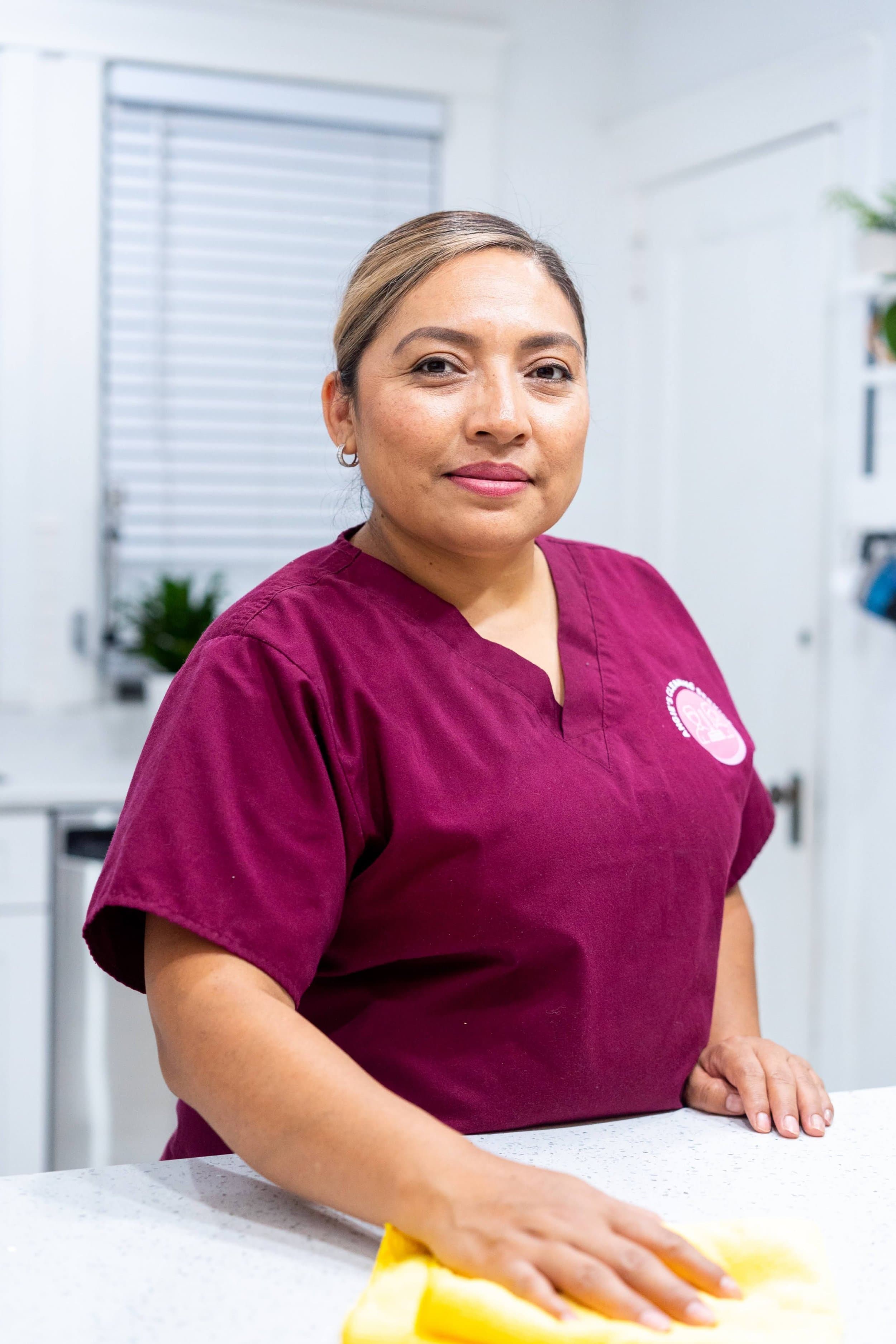 Angie in a maroon scrub top at a bright white countertop, cloth in hand.